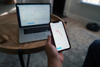 A person holds a smartphone displaying a stock market trading app with candlestick charts. A laptop is on a wooden table with a similar financial chart, indicating active trading or analysis. The setting suggests a home office or casual workspace.
