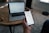 A person holds a smartphone displaying a stock market trading app with candlestick charts. A laptop is on a wooden table with a similar financial chart, indicating active trading or analysis. The setting suggests a home office or casual workspace.