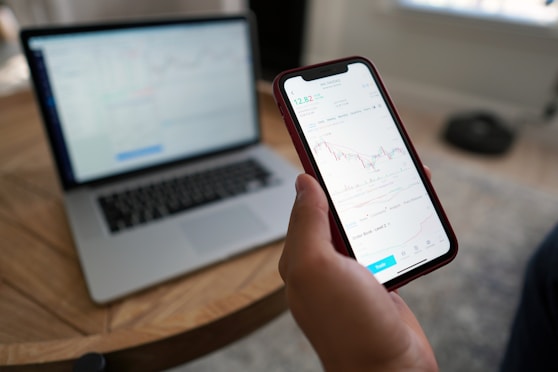 A hand holding a smartphone displaying a stock trading chart with details on stock prices. The phone is above a wooden table where a blurred laptop screen in the background shows similar financial data. Natural light comes in from the window, creating a relaxed working environment.