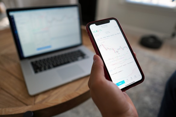 A hand holding a smartphone displaying a stock trading chart with details on stock prices. The phone is above a wooden table where a blurred laptop screen in the background shows similar financial data. Natural light comes in from the window, creating a relaxed working environment.
