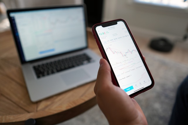 A hand holding a smartphone displaying a stock trading chart with details on stock prices. The phone is above a wooden table where a blurred laptop screen in the background shows similar financial data. Natural light comes in from the window, creating a relaxed working environment.