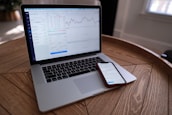 A laptop and smartphone are placed on a wooden table. Both devices display financial charts and data, suggesting involvement in stock trading or market analysis. The setting appears to be indoors with natural light coming through a window.