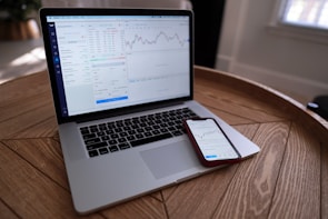 A laptop and smartphone are placed on a wooden table. Both devices display financial charts and data, suggesting involvement in stock trading or market analysis. The setting appears to be indoors with natural light coming through a window.