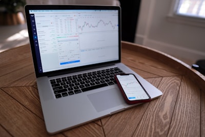 A laptop and smartphone are placed on a wooden table. Both devices display financial charts and data, suggesting involvement in stock trading or market analysis. The setting appears to be indoors with natural light coming through a window.