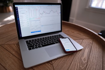 A laptop and smartphone are placed on a wooden table. Both devices display financial charts and data, suggesting involvement in stock trading or market analysis. The setting appears to be indoors with natural light coming through a window.