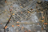 A detailed scale model of an ancient city or architectural plan is surrounded by scattered leaves. The model is crafted with precision, featuring buildings, roads, and open spaces. The material appears metallic, with a worn, weathered surface.