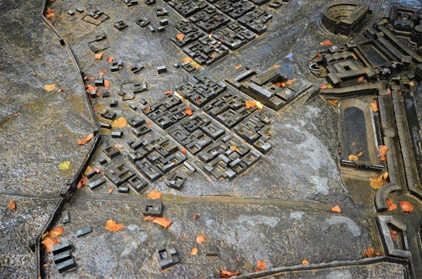 A detailed scale model of an ancient city or architectural plan is surrounded by scattered leaves. The model is crafted with precision, featuring buildings, roads, and open spaces. The material appears metallic, with a worn, weathered surface.