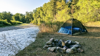 A serene campsite features a blue tent set up near a gently flowing river, surrounded by lush greenery and trees. In the foreground, there is a fire pit constructed from a circle of stones on the earthy ground.