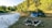 A serene campsite features a blue tent set up near a gently flowing river, surrounded by lush greenery and trees. In the foreground, there is a fire pit constructed from a circle of stones on the earthy ground.