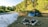 A serene campsite features a blue tent set up near a gently flowing river, surrounded by lush greenery and trees. In the foreground, there is a fire pit constructed from a circle of stones on the earthy ground.