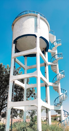 A prominent water tower with a cylindrical tank supported by a white, lattice-like framework stands tall against a bright blue sky. The structure includes an external spiral staircase with blue railings, allowing access to the top of the tower. Surrounding the base are some green shrubs and trees, adding a touch of nature to the scene.