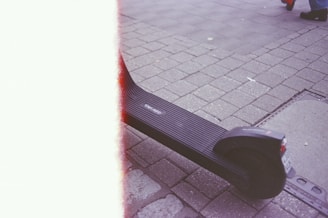 A scooter is parked on a cobblestone street, with a visible license plate and the word 'tier.app' on the footboard. The left side of the image has a light leak or film burn effect. On the top right corner, parts of a person's legs in blue pants and dark shoes are seen.