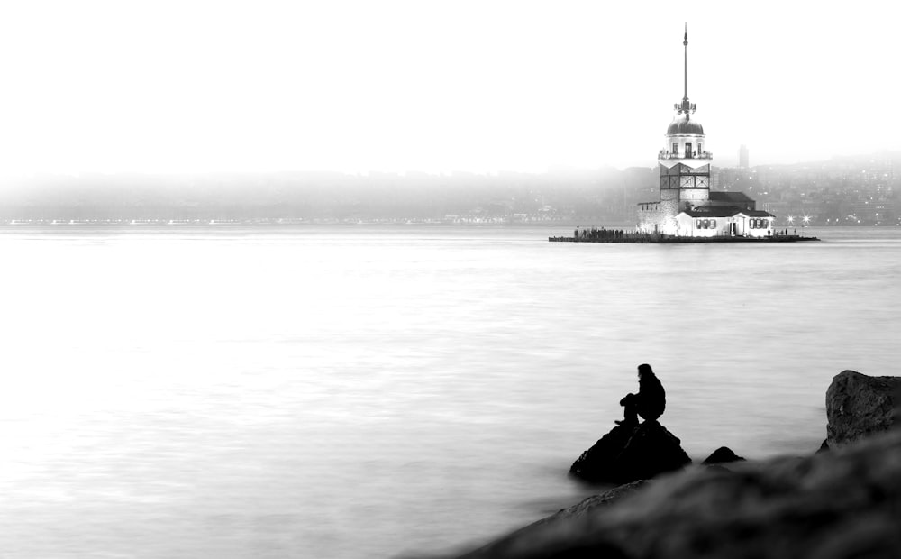 grayscale photo of man sitting on rock near body of water