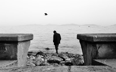 man in black jacket standing on rock near body of water during daytime contemplative zoom background