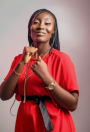 A person wearing a bright red dress with a waist belt and accessorized with a gold watch and chain. The individual has braided hair and displays a confident posture with clenched fists.