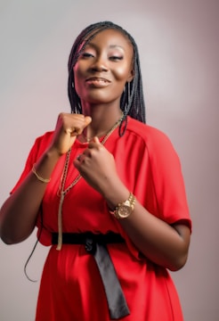 A person wearing a bright red dress with a waist belt and accessorized with a gold watch and chain. The individual has braided hair and displays a confident posture with clenched fists.