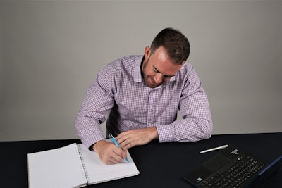 A person in a checkered shirt is sitting at a table, writing in a notebook with a pen. A laptop is placed on the table beside the notebook.