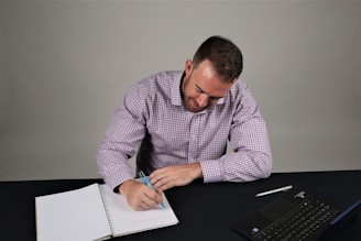 A person in a checkered shirt is sitting at a table, writing in a notebook with a pen. A laptop is placed on the table beside the notebook.