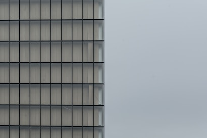A minimalist architectural photograph featuring the side of a modern building with a grid-like pattern of windows. The right half of the image is dominated by a plain, light-gray sky, creating a stark contrast between the structured lines of the building and the open space.