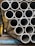 A stack of metallic tubes arranged in a horizontal pattern, with the ends of the tubes facing the viewer. The tubes have a slightly worn appearance and are resting on a blue metal support. The scene appears to be in an industrial setting with various shades of metal and other structures in the background.