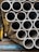 A stack of metallic tubes arranged in a horizontal pattern, with the ends of the tubes facing the viewer. The tubes have a slightly worn appearance and are resting on a blue metal support. The scene appears to be in an industrial setting with various shades of metal and other structures in the background.