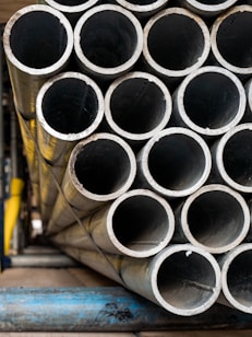 A stack of metallic tubes arranged in a horizontal pattern, with the ends of the tubes facing the viewer. The tubes have a slightly worn appearance and are resting on a blue metal support. The scene appears to be in an industrial setting with various shades of metal and other structures in the background.