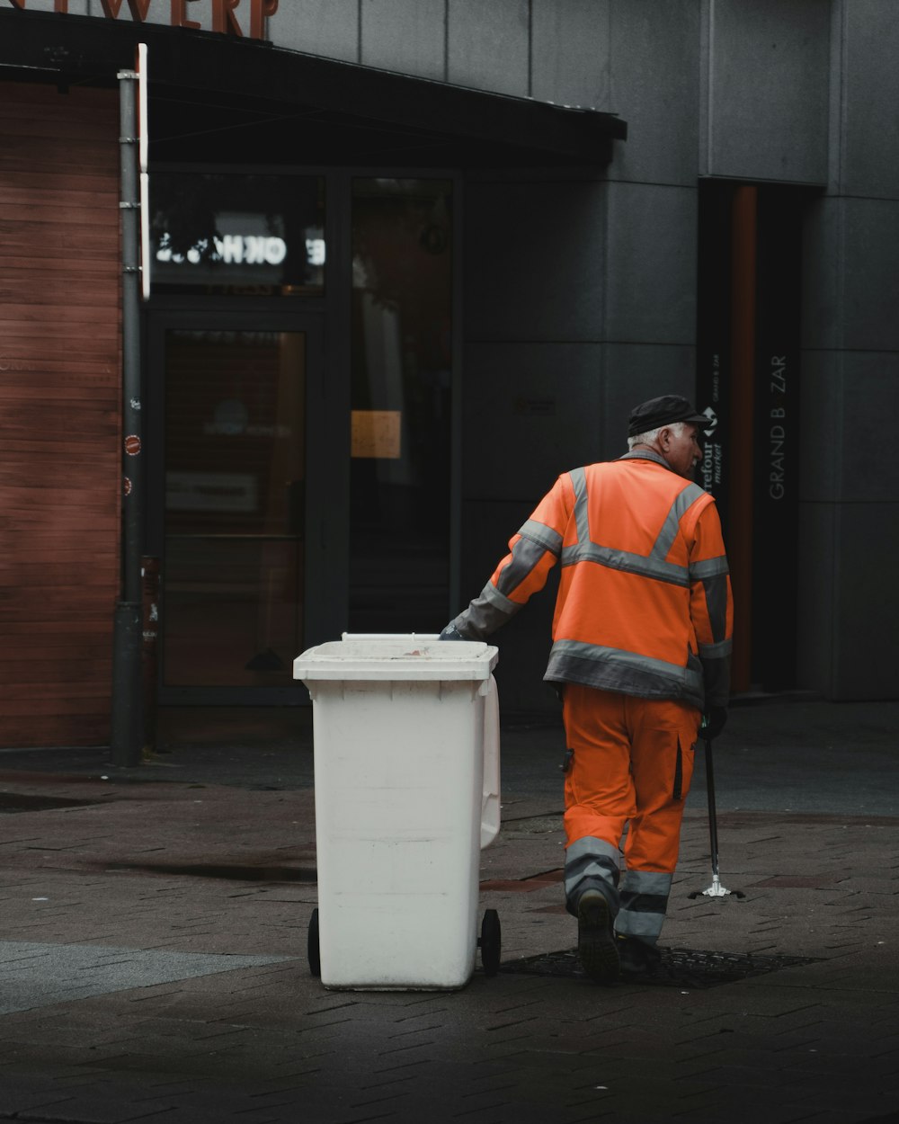 オレンジ色のジャケットと黒いズボンを着た男性が白いプラスチックのゴミ箱のそばに立っています