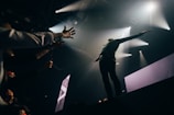 A performer is on stage with arms outstretched, engaging with an enthusiastic audience reaching out from the left. Bright spotlights shine above, creating a dramatic atmosphere.