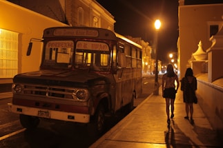 An old bus is parked on a dimly lit street at night, with two people walking along the sidewalk in the foreground. The street is illuminated by a series of streetlamps, casting a warm glow on the surrounding buildings. The atmosphere suggests a quiet, possibly residential area.