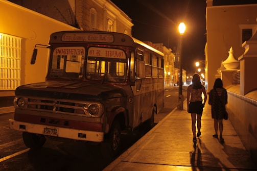 An old bus is parked on a dimly lit street at night, with two people walking along the sidewalk in the foreground. The street is illuminated by a series of streetlamps, casting a warm glow on the surrounding buildings. The atmosphere suggests a quiet, possibly residential area.
