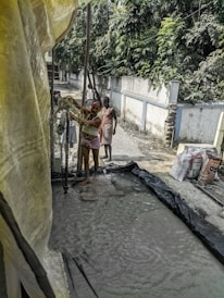 A group of people is engaged in a manual task, standing on wet ground surrounded by a makeshift setup with plastic sheets. A man in the foreground is handling some equipment, while others stand nearby. Bags of materials are visible, suggesting construction or manual labor work. Dense greenery lines the wall in the background.