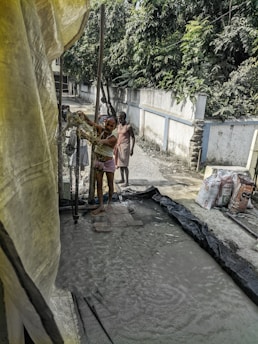 A group of people is engaged in a manual task, standing on wet ground surrounded by a makeshift setup with plastic sheets. A man in the foreground is handling some equipment, while others stand nearby. Bags of materials are visible, suggesting construction or manual labor work. Dense greenery lines the wall in the background.