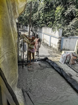 A group of people is engaged in a manual task, standing on wet ground surrounded by a makeshift setup with plastic sheets. A man in the foreground is handling some equipment, while others stand nearby. Bags of materials are visible, suggesting construction or manual labor work. Dense greenery lines the wall in the background.