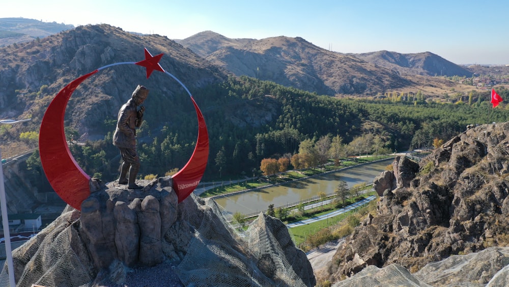 Hombre con chaqueta y pantalones negros sosteniendo la bandera roja de pie en la roca cerca del lago durante el día