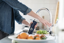 A person in a denim shirt is holding a navy blue water bottle under a running tap in a kitchen setting. Fresh fruits and vegetables are visible in the foreground.