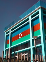 A modern building facade featuring a prominent display of the Azerbaijani flag, characterized by blue, red, and green horizontal stripes, along with a crescent and an eight-pointed star. The structure incorporates glass and metal elements with a sleek, contemporary design. A streetlamp and fencing are visible in front of the building, with some trees in the lower foreground.