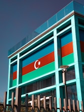 A modern building facade featuring a prominent display of the Azerbaijani flag, characterized by blue, red, and green horizontal stripes, along with a crescent and an eight-pointed star. The structure incorporates glass and metal elements with a sleek, contemporary design. A streetlamp and fencing are visible in front of the building, with some trees in the lower foreground.