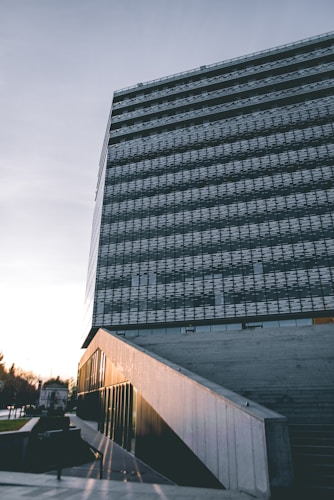 A modern architectural structure with a large glass facade. The building has clean lines and a geometric design. The sky appears slightly overcast, while the sunlight reflects off the glass panels, creating a warm glow. The foreground includes a concrete staircase leading to the building's entrance.
