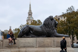 A large stone statue of a lion is prominently displayed on a plinth in an open public area. Two people are sitting on the ledge beside the lion, smiling toward the camera. In the background, a tall, white clock tower rises above lush green trees, adding to the urban landscape setting.