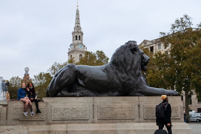 A large stone statue of a lion is prominently displayed on a plinth in an open public area. Two people are sitting on the ledge beside the lion, smiling toward the camera. In the background, a tall, white clock tower rises above lush green trees, adding to the urban landscape setting.