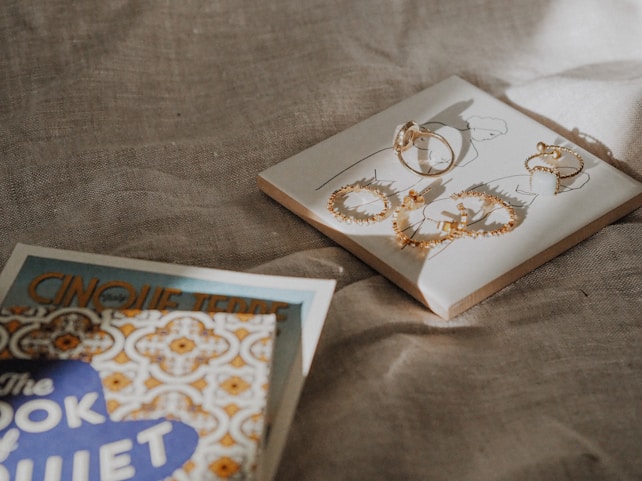 On a textured fabric surface, a square card holds several gold hoop earrings, each adorned with small stones. Adjacent to this card, a partially visible book titled 'The Book of Quiet' and another with 'Cinque Terre' on its cover, show intricate patterns and lettering.