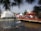 A lively outdoor event featuring a fountain with water jets in the foreground. People are gathering around red and white tents with vibrant decorations. A red bridge crosses over a pond, adding a festive atmosphere.