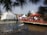 A lively outdoor event featuring a fountain with water jets in the foreground. People are gathering around red and white tents with vibrant decorations. A red bridge crosses over a pond, adding a festive atmosphere.