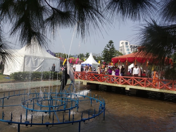 A lively outdoor event featuring a fountain with water jets in the foreground. People are gathering around red and white tents with vibrant decorations. A red bridge crosses over a pond, adding a festive atmosphere.