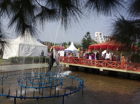 A lively outdoor event featuring a fountain with water jets in the foreground. People are gathering around red and white tents with vibrant decorations. A red bridge crosses over a pond, adding a festive atmosphere.
