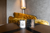 A white mug with a black cannabis leaf design is placed on a wooden table next to a stylish perfume box. In the background, a luxurious, mustard-yellow velvet sofa with a curved back and soft cushions is accompanied by a tall, modern lamp against a textured wall.