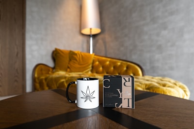 A white mug with a black cannabis leaf design is placed on a wooden table next to a stylish perfume box. In the background, a luxurious, mustard-yellow velvet sofa with a curved back and soft cushions is accompanied by a tall, modern lamp against a textured wall.