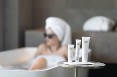 A serene spa setting with three beauty product tubes displayed prominently on a small round table. In the background, a person relaxing in a bathtub with a towel wrapped around their head and sunglasses.