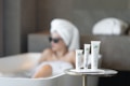 A serene spa setting with three beauty product tubes displayed prominently on a small round table. In the background, a person relaxing in a bathtub with a towel wrapped around their head and sunglasses.
