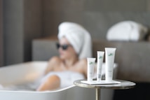A serene spa setting with three beauty product tubes displayed prominently on a small round table. In the background, a person relaxing in a bathtub with a towel wrapped around their head and sunglasses.